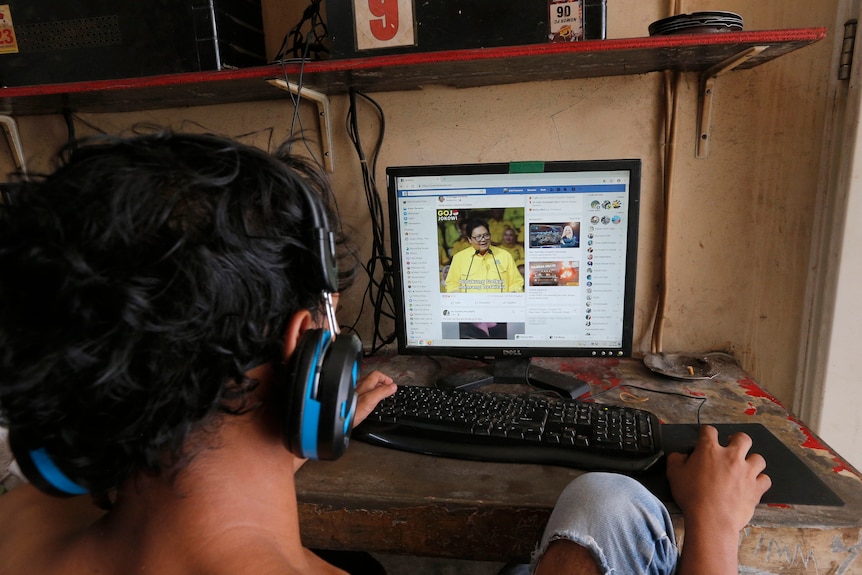 The back of a younng Indonesian person's head, wearing headphones while browsing Facebook. 