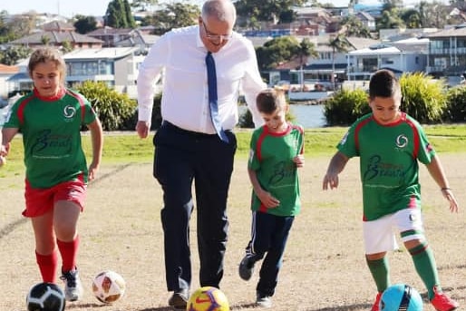 Scott Morrison dribbles a soccer ball alongside children on a suburban soccer field.