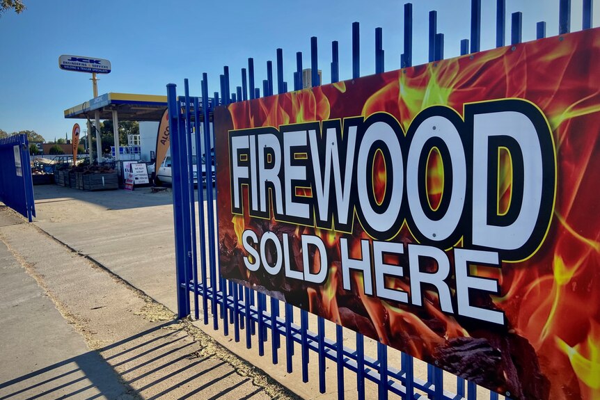 'Firewood sold here' sign tied to a blue fence with a yellow and blue store called JCK Engineering & Services in background