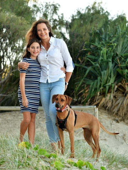 A woman stands with a young girl and dog on a sand dune.