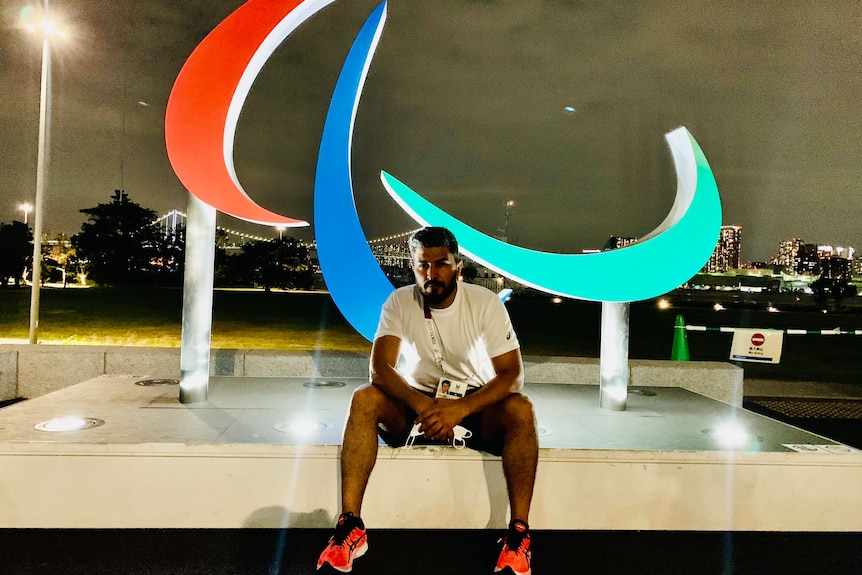 A young middle eastern man in white T-shirt and shorts sits in front of colourfully lit display at night.