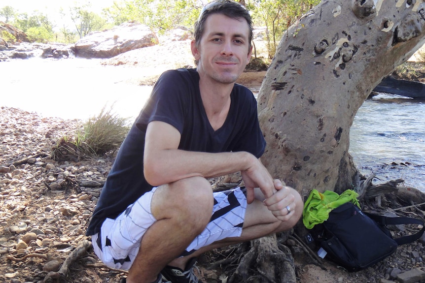 A man kneels down next to a tree and river posing for a photo in shorts and a t-shirt.