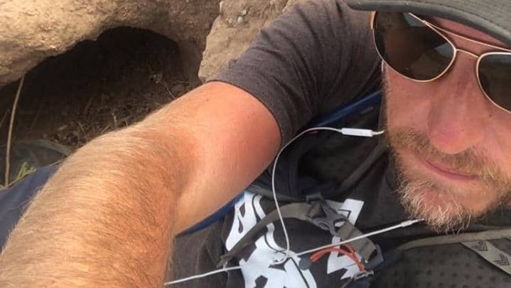 A man in a Nike visor cap and aviator sunglasses sits in a large wombat burrow.