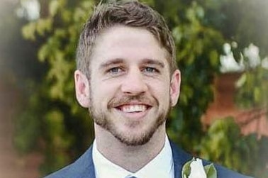 A smiling young man with dark hair and a neat beard wearing groomsman-style suit.