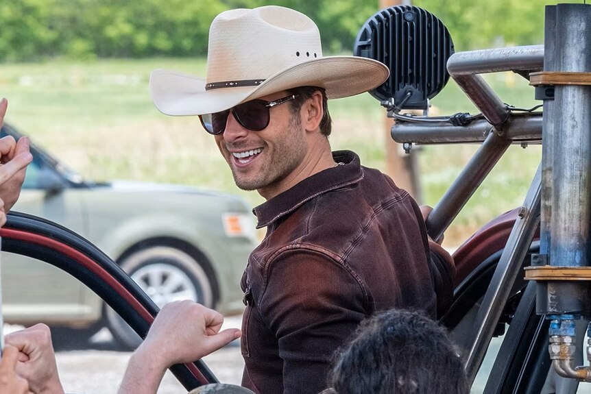 A handsome man in a cowboy hat and sunglasses grins back at a cheering crowd as he climbs into a vehicle.