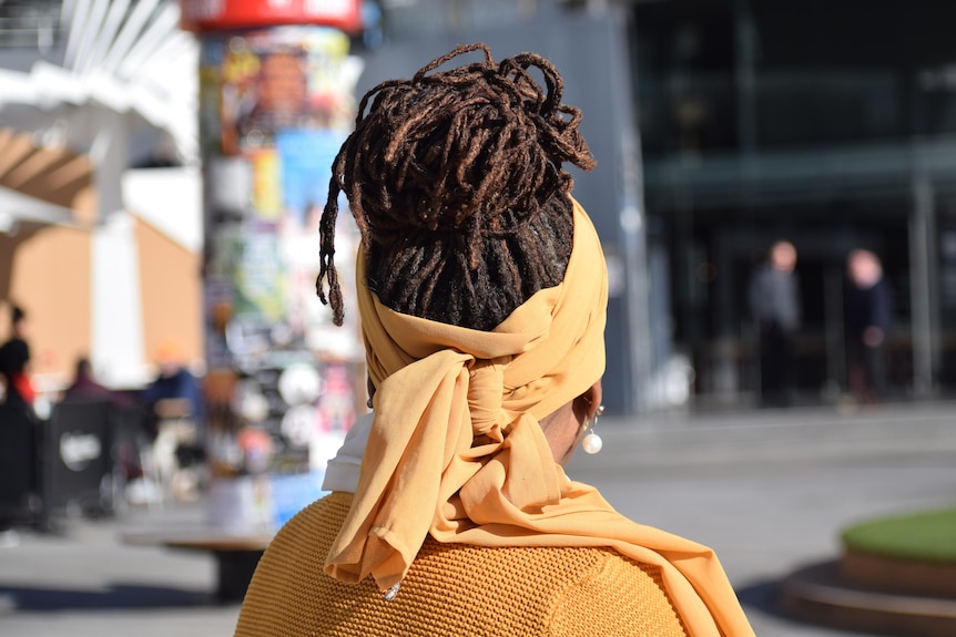 An image showing the back of a woman's head. Her hair is in dreadlocks and tied up. She is wearing yellow.