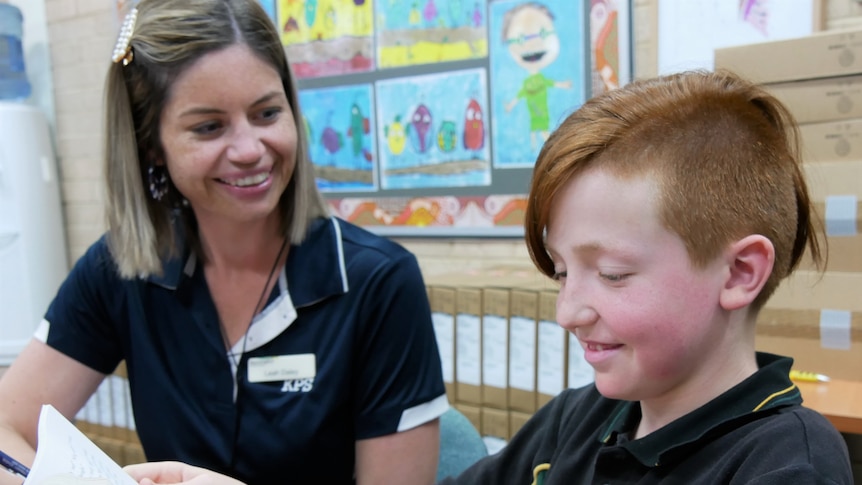 A young woman with short blonde hair smiles at a red-headed boy as he learns to read.