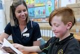 A young woman with short blonde hair smiles at a red-headed boy as he learns to read.