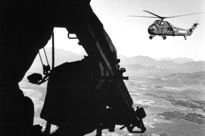 Black and white photo of soldier inside a helicopter looking at another helicopter in air.