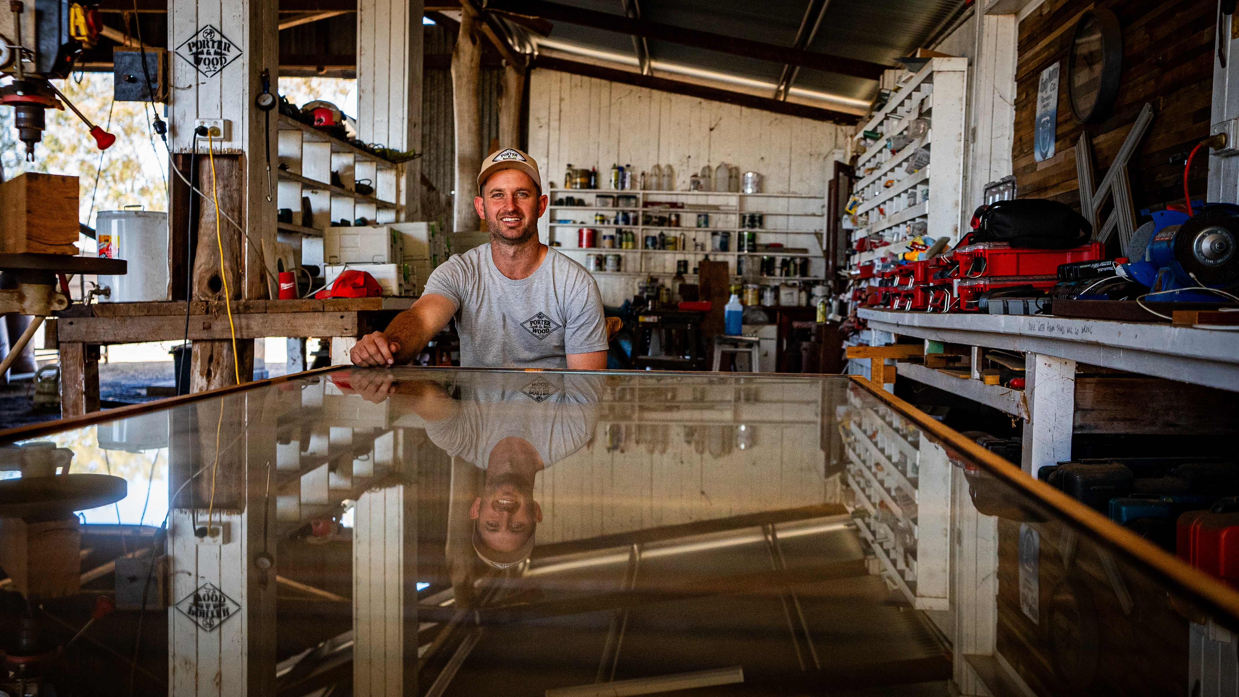 A man sits behind his long working table in his workshop filled with multiple tools. 