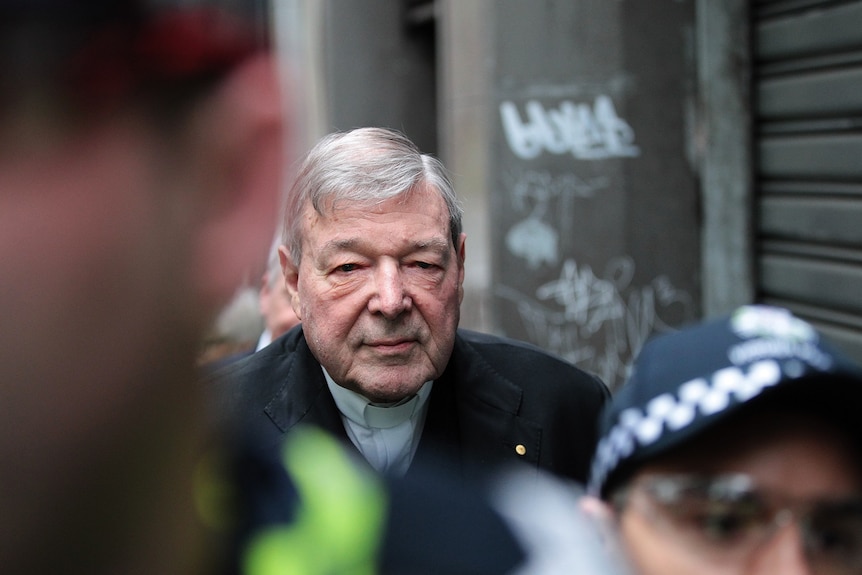 George Pell walking in an alley way with police officers out of focus in front of him.