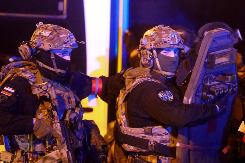 Two armed men in uniform, with their faces covered, look on.