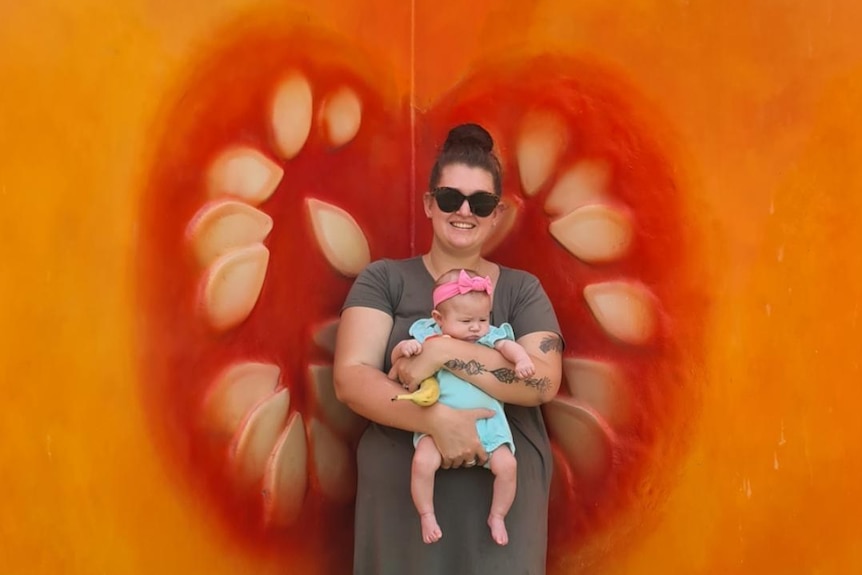 A woman holds a baby while standing in front of a large pumpkin.