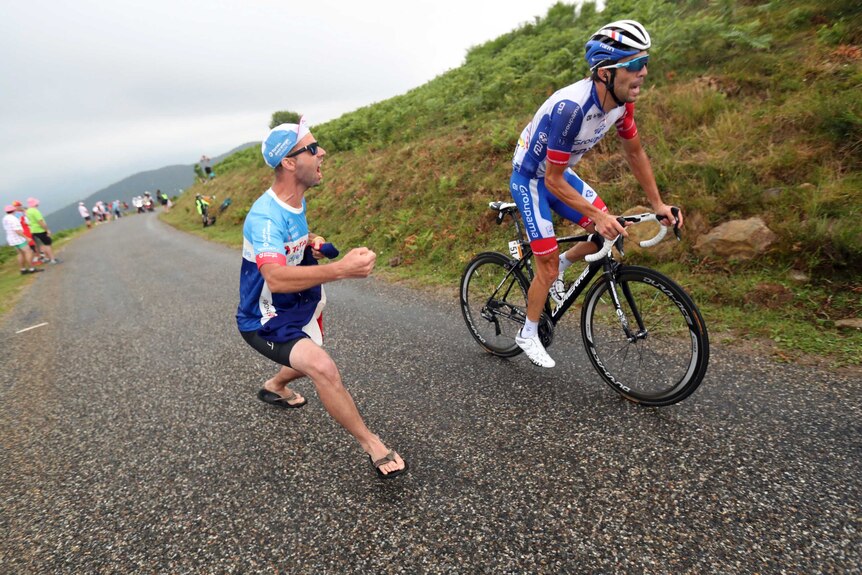 A fan holding a French flag runs on the road and cheers on Thibaut Pinot, who is cycling up a hill.