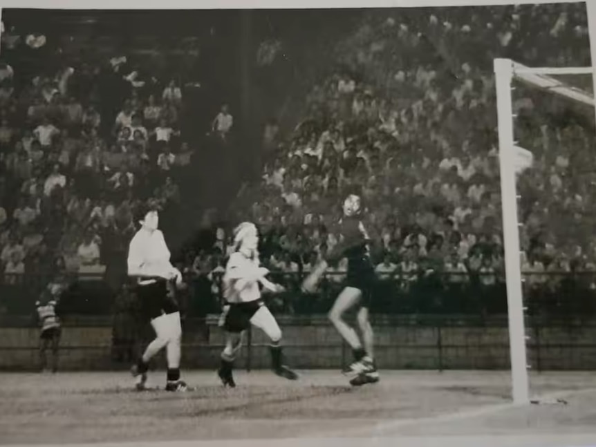 An old photograph showing a soccer player wearing a light coloured jersey scoring a goal during a match in front of a crowd