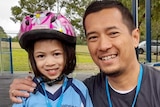 A girl wearing a bike helmet smiles next to her bike and her dad.