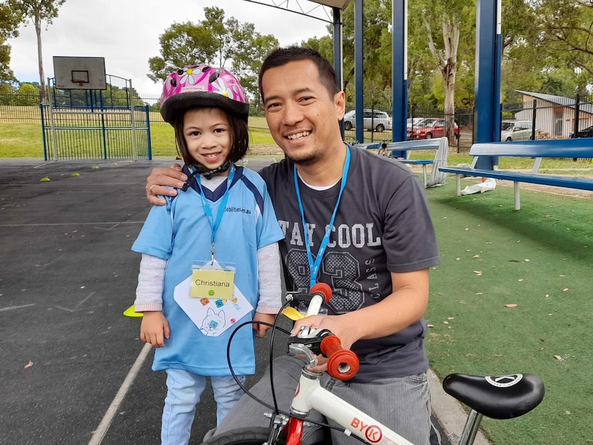 A girl wearing a bike helmet smiles next to her bike and her dad.