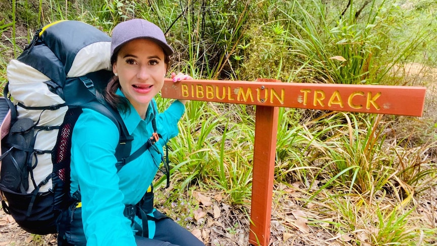 A young woman in hiking gear squats down next to a sign that reads Bibbulmun Track