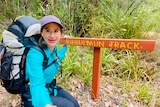 A young woman in hiking gear squats down next to a sign that reads Bibbulmun Track