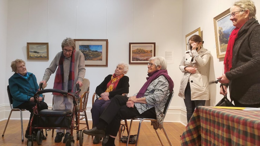 A group of elderly women sitting in a semi-circle with paintings on the walls behind them.