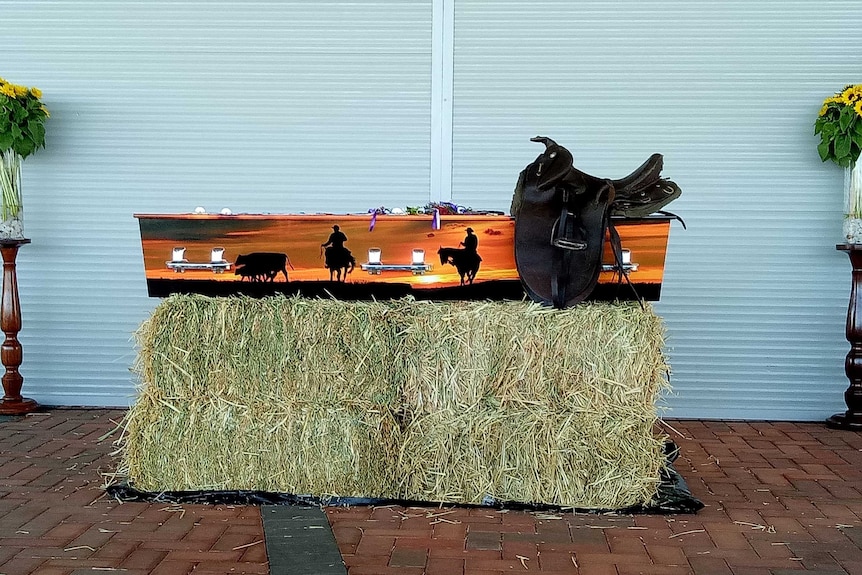 A coffin with images of stock riders and cattle, sitting on hay bales with saddle on top.