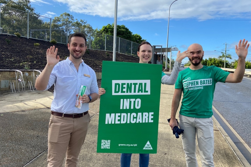 Greens' candidate for the seat of Brisbane Stephen Bates  waves to camera with two supporters with a sign 'Dental into Medicare'