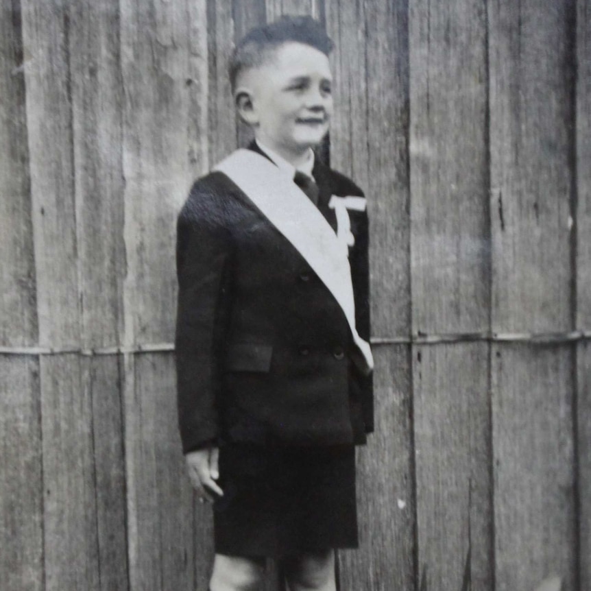 A young boy standing in front of a wooden fence dressed in a suit with a white sash