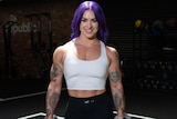 A woman with purple hair and strong arms smiles with weights in both hands.