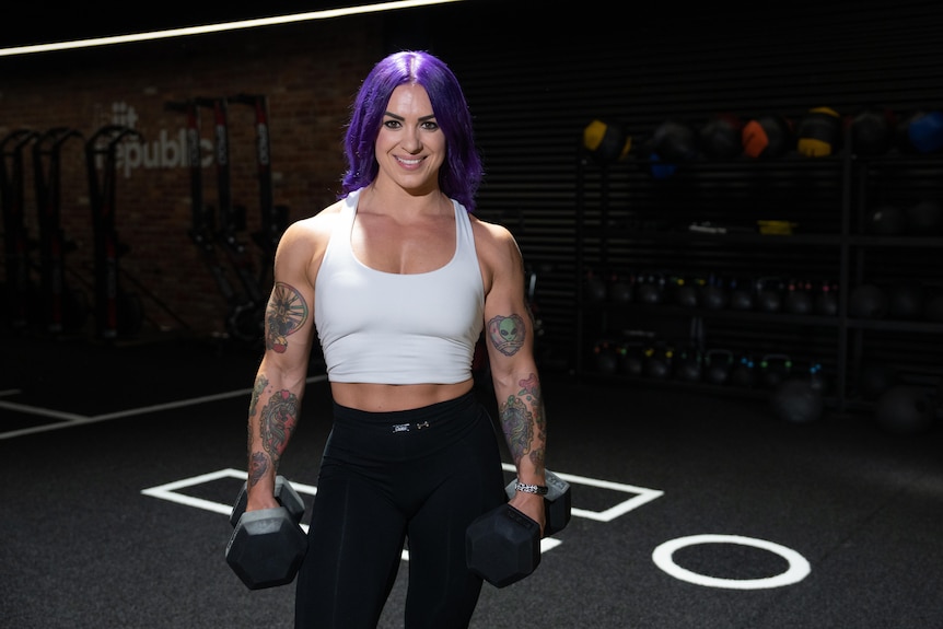 A woman with purple hair and strong arms smiles with weights in both hands.