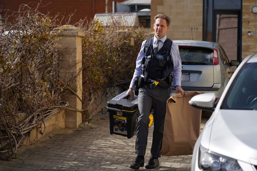 A police officer in plain clothes and a police vest carries a large paper bag and a plastic container from a building near cars.