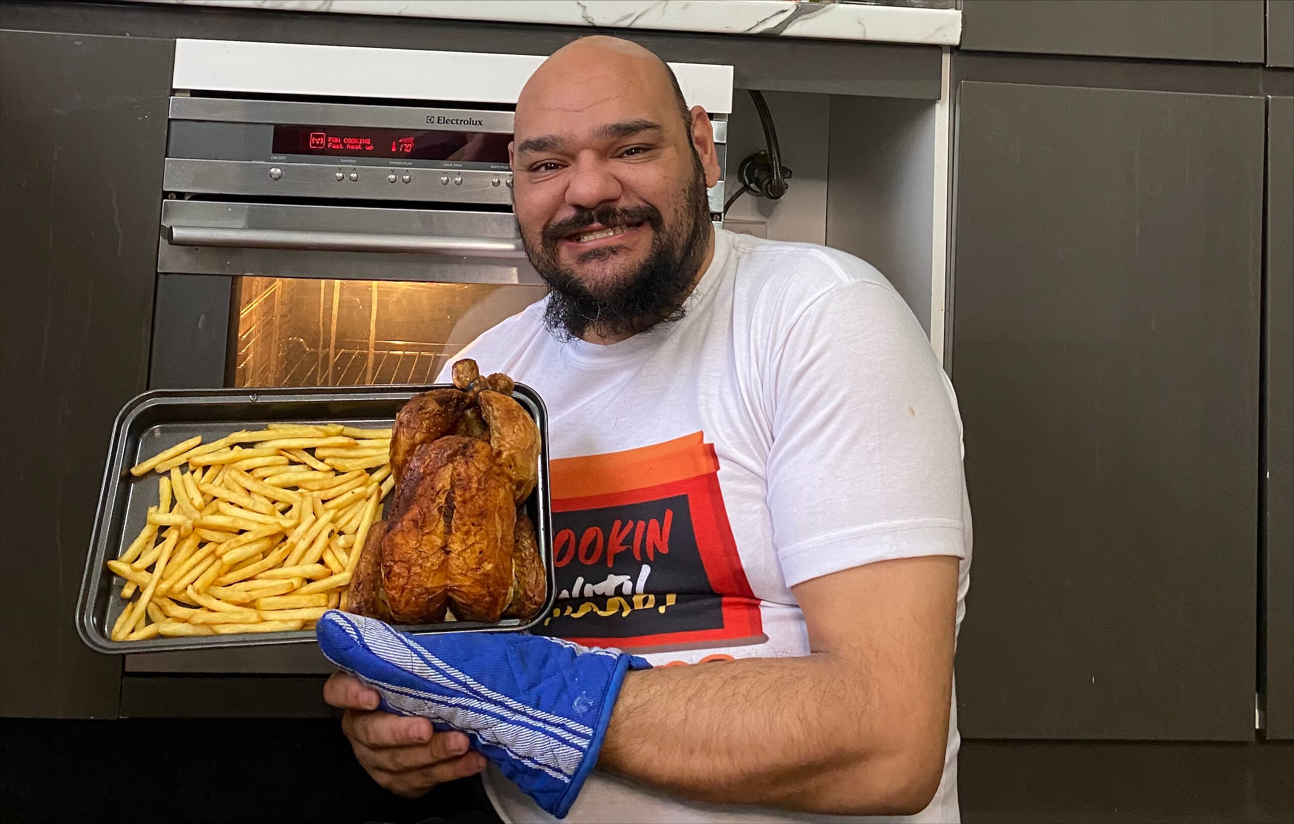 Nathan grins, holding up an oven tray of chicken and chips.