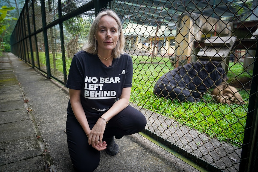 Woman in a black shirt that reads 'No bear left behind' kneeling next to a bear in an enclosure.
