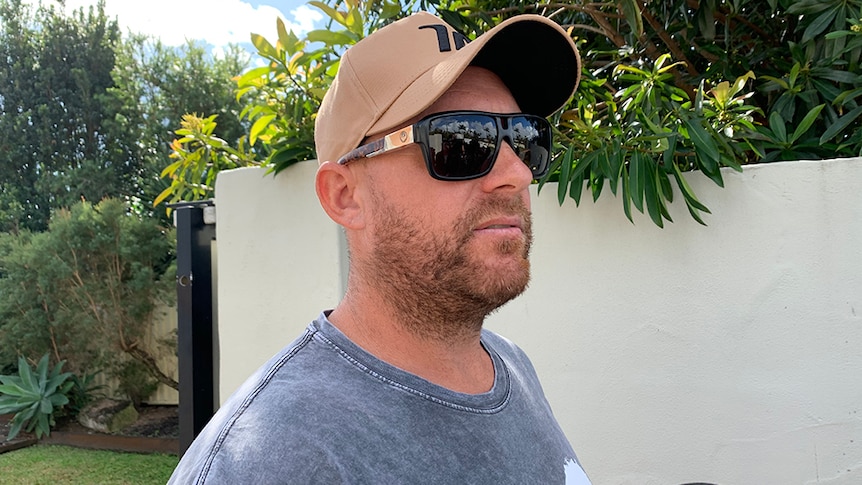 Man with  dark sunglasses and brown hat stands outside a white fence with a microphone under his chin