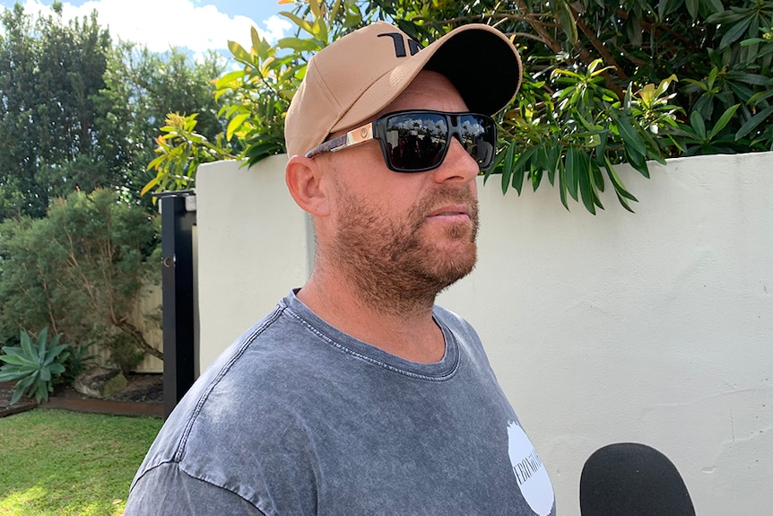 Man with  dark sunglasses and brown hat stands outside a white fence with a microphone under his chin