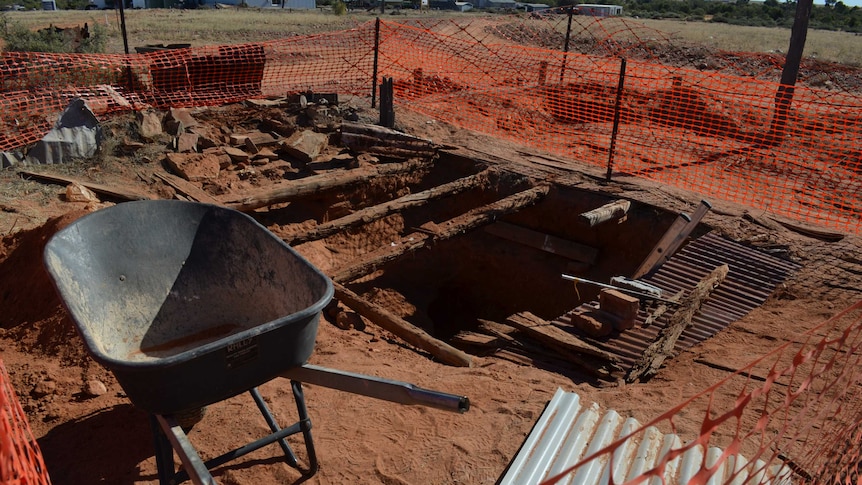 A fenced off area shows a large hole in the ground with rotten wooden beams across it and a wheelbarrow nearby.