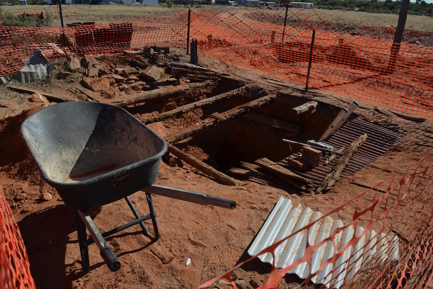 A fenced off area shows a large hole in the ground with rotten wooden beams across it and a wheelbarrow nearby.