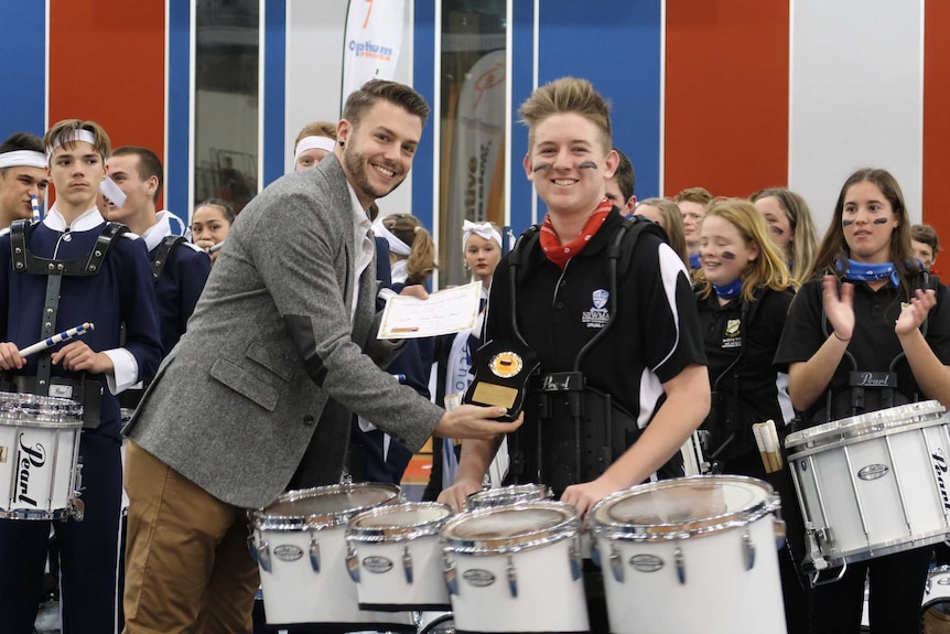 A man smiles wearing a grey tweed coat and hands an award to a student, in front of many students with percussive instruments.