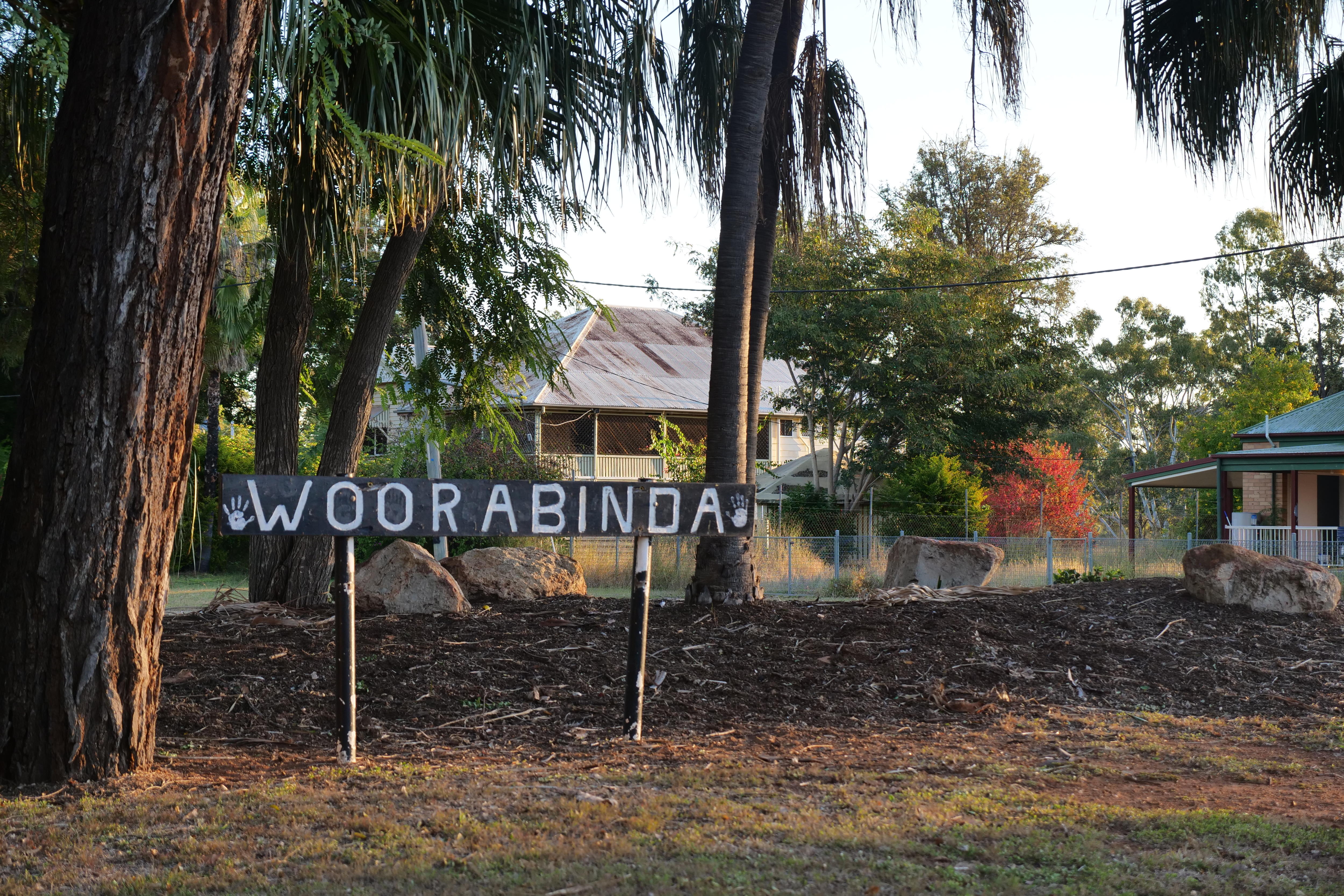 A sign in a garden with trees and houses behind it reads Woorabinda.