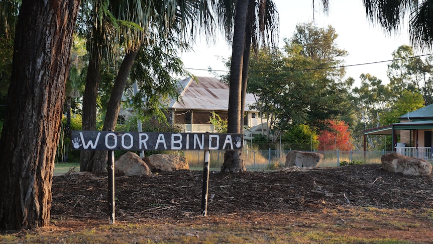 A sign in a garden with trees and houses behind it reads Woorabinda.