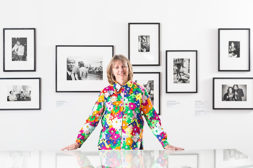 A 60-year-old woman with long bob haircut in a colourful floral shirt stands in front of black and white photos hung on a wall