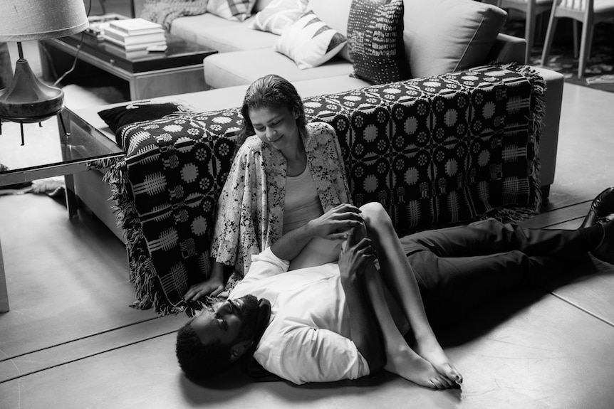 Photo of young man and woman on lounge room floor, him supine and her sitting against couch with her legs over his torso.
