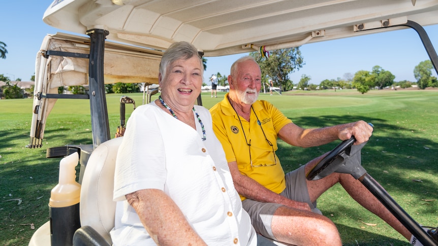 Two elderly people sit in a golf buggy smiling on a sunny day. 