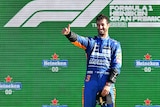 Race car driver wearing blue stands on the winner's podium and gives a thumbs-up.