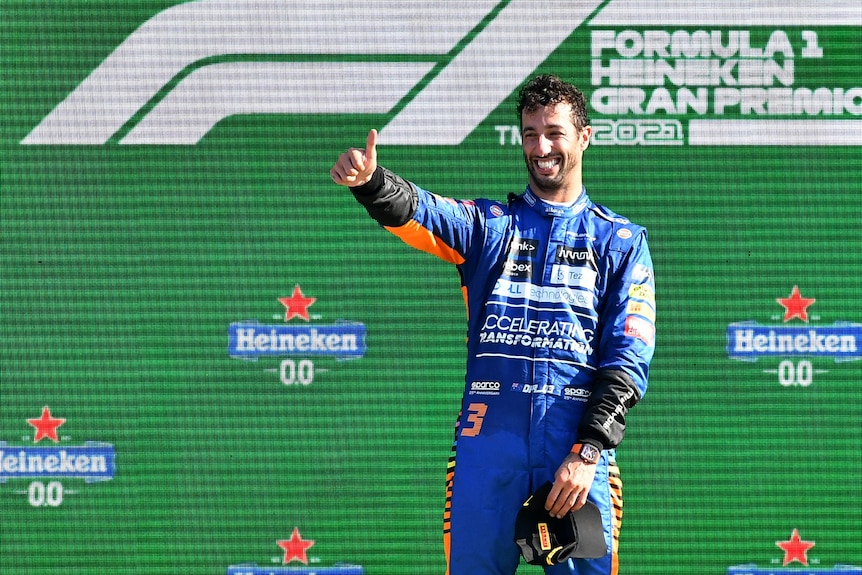 Race car driver wearing blue stands on the winner's podium and gives a thumbs-up.