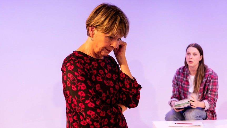 50ish woman with short hair and black & red blouse upstage staring down looking worried; 20ish girl sits on chair downstage.