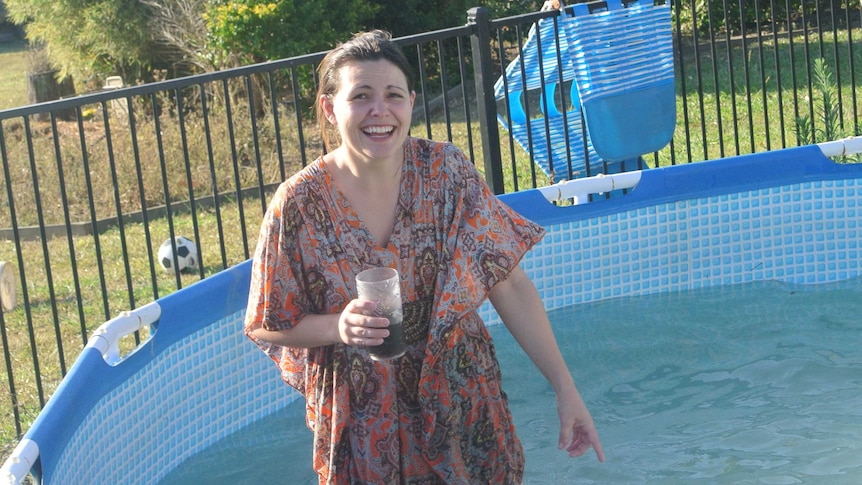 A woman wearing a flowy kaftan dress laughs while standing in a backyard wading pool.