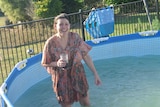 A woman wearing a flowy kaftan dress laughs while standing in a backyard wading pool