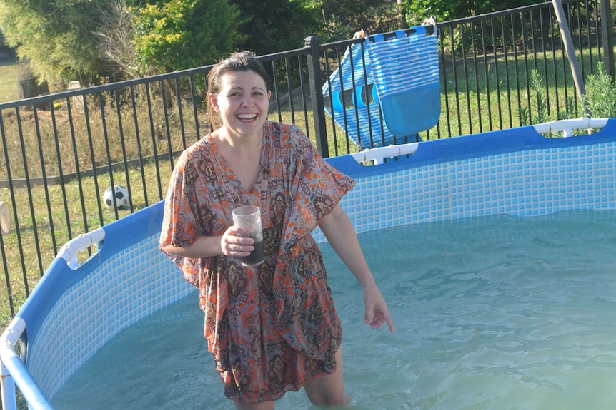 A woman wearing a flowy kaftan dress laughs while standing in a backyard wading pool