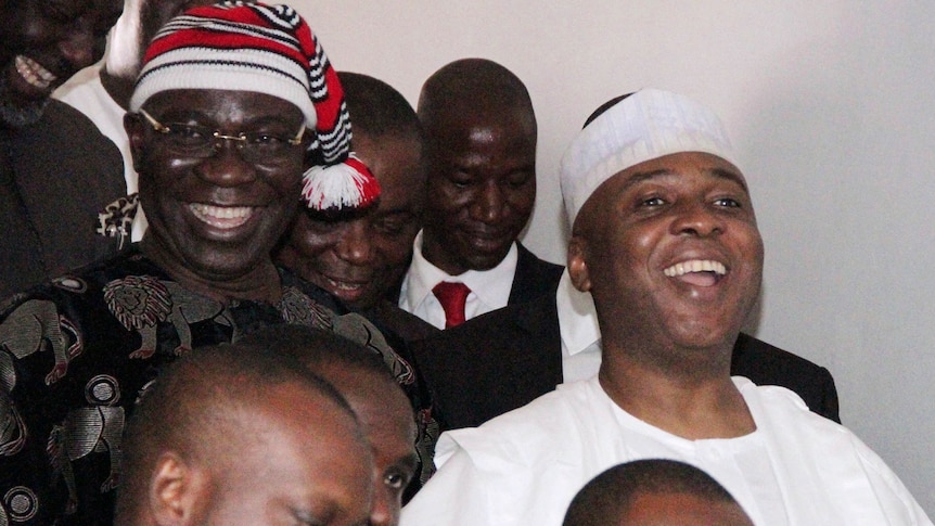 Two smiling men in hats stand among a group of other men.
