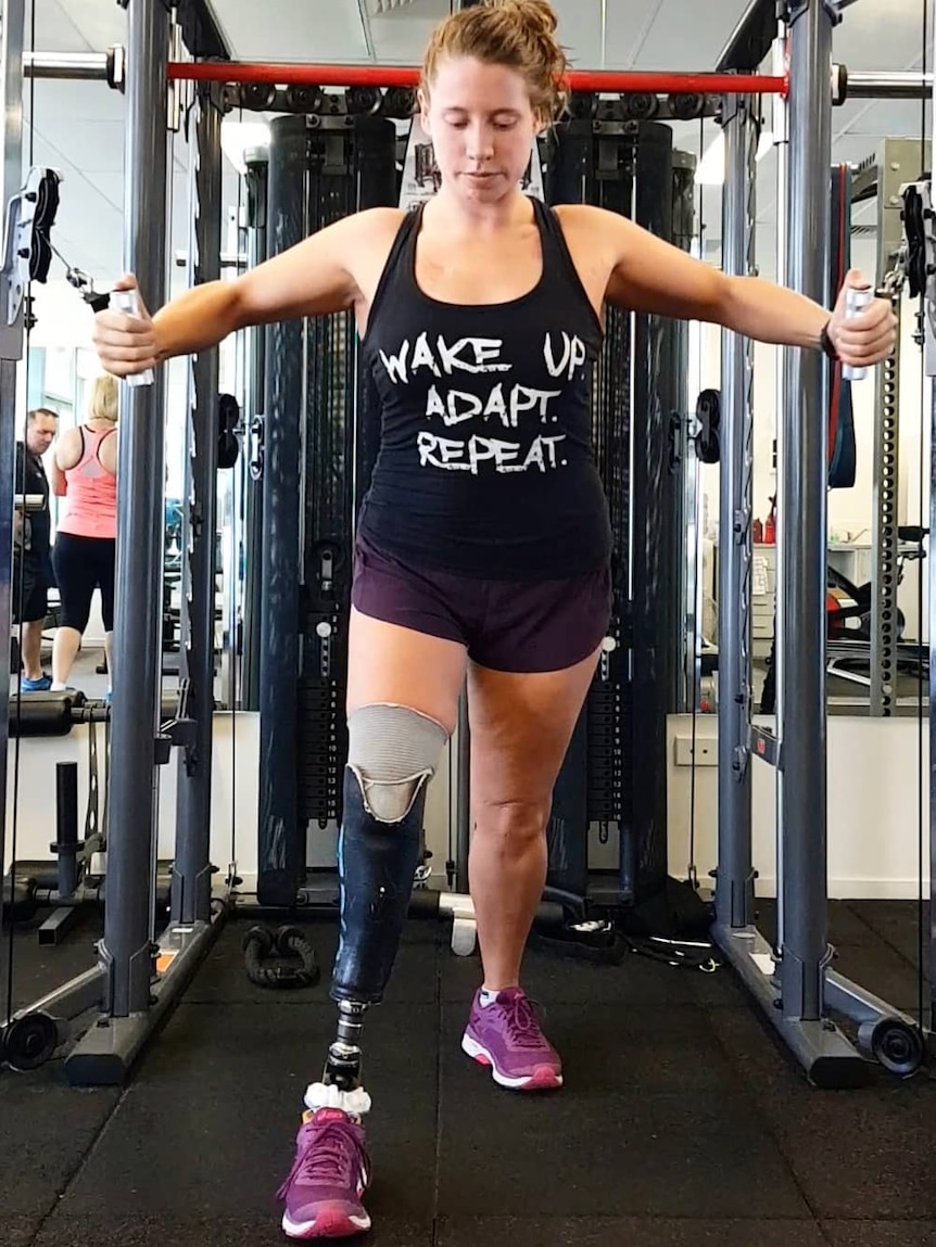 A woman lifts weights at a gym on a machine, while wearing a black singlet. Her right leg is amputated below the knee.